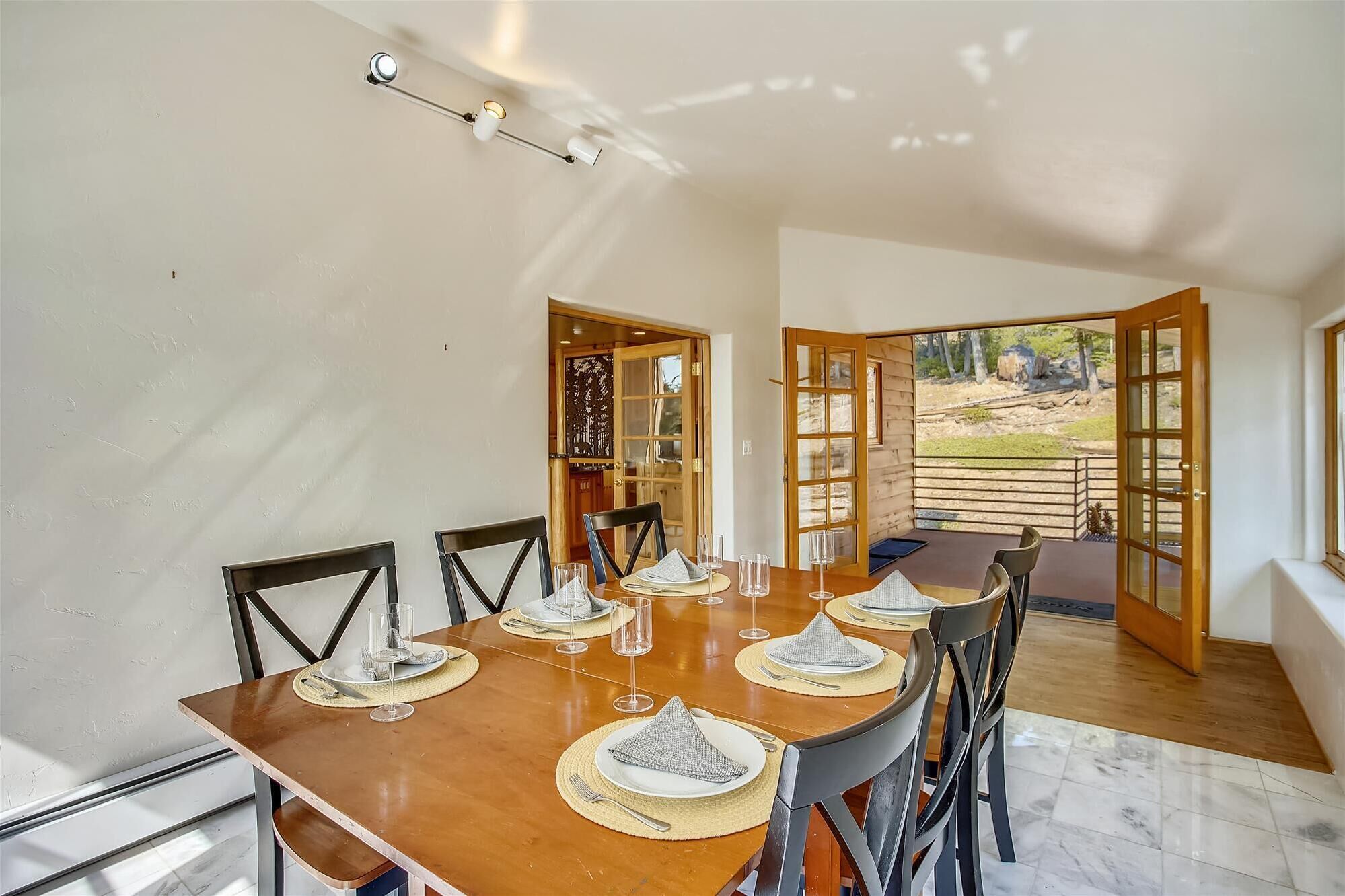 A dining area with a wooden table set for six, surrounded by chairs. There are two doors leading to an outdoor area.