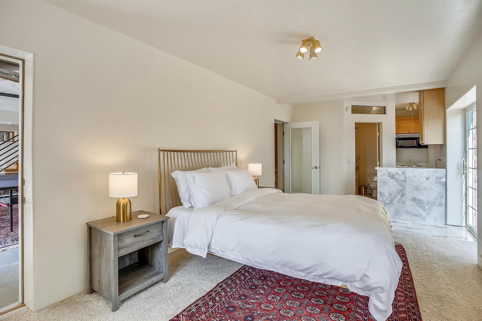 A bedroom with a bed, nightstands, a red patterned rug, a ceiling light fixture, and a view of the bathroom.