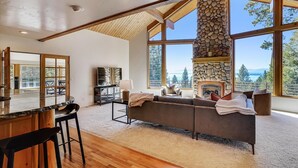 A spacious living room with a stone fireplace, large windows offering a view of the outdoors, and a cozy seating area. - Modern, Hot Tub, Lake View, Short Drive to Beach, Restaurants, and Shops- Flying Flamingo (Kings Beach)