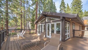 Een houten terras met lounge stoelen, een gebouw met glazen wanden en hoge bomen op de achtergrond.