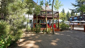 A wooden cabin with a deck, surrounded by pine trees and a clear sky. - Sandy yard that backs to the Beach! Walk to town -Lakefront on the Beach (Kings Beach)