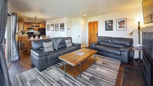 A modern living room with two black leather sofas, a wooden coffee table, and a striped rug. There is a dining area with wooden chairs and a table, and a kitchen in the background. - Sandy yard that backs to the Beach! Walk to town -Lakefront on the Beach (Kings Beach)