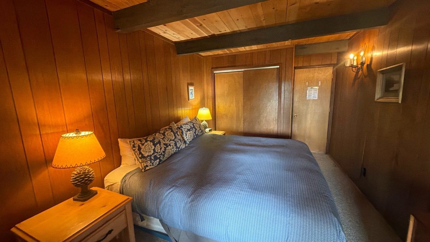 A wooden-paneled room with a bed, a nightstand with a lamp, and a door with a sign.