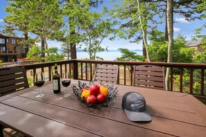 Una terraza de madera con una mesa dispuesta para dos personas, que incluye una botella de vino, dos copas de vino y una cesta de frutas. También se observa un tapón en la mesa.