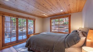 Un dormitorio con techo de madera, ventanas amplias, una cama con ropa de cama gris y una mesita de noche con lámpara.