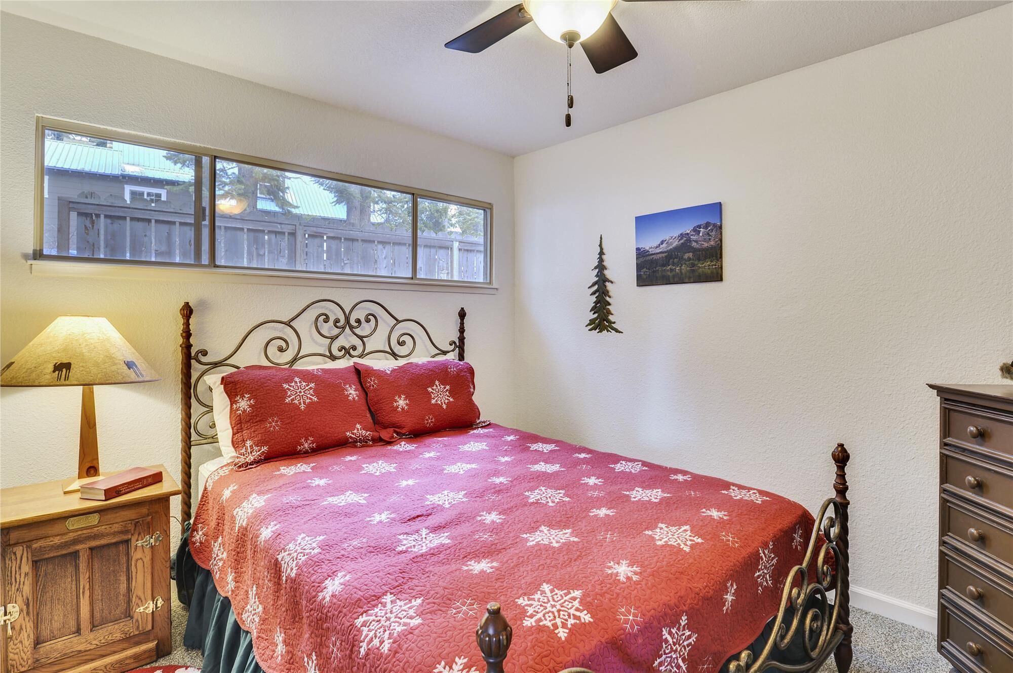 Une chambre à coucher comprenant un lit, une table de chevet en bois, une lampe, une commode, une fenêtre et un ventilateur de plafond.