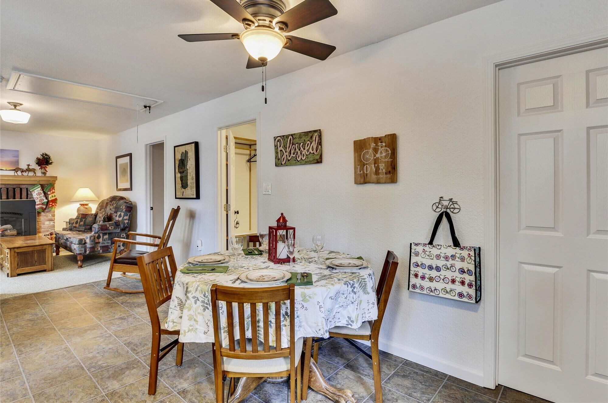 Une salle à manger avec une table dressée pour quatre personnes, entourée de chaises en bois. Un ventilateur de plafond est visible au-dessus de la table, et les murs sont décorés, notamment avec un panneau portant l’inscription « Béni » et une œuvre sur le thème du vélo.