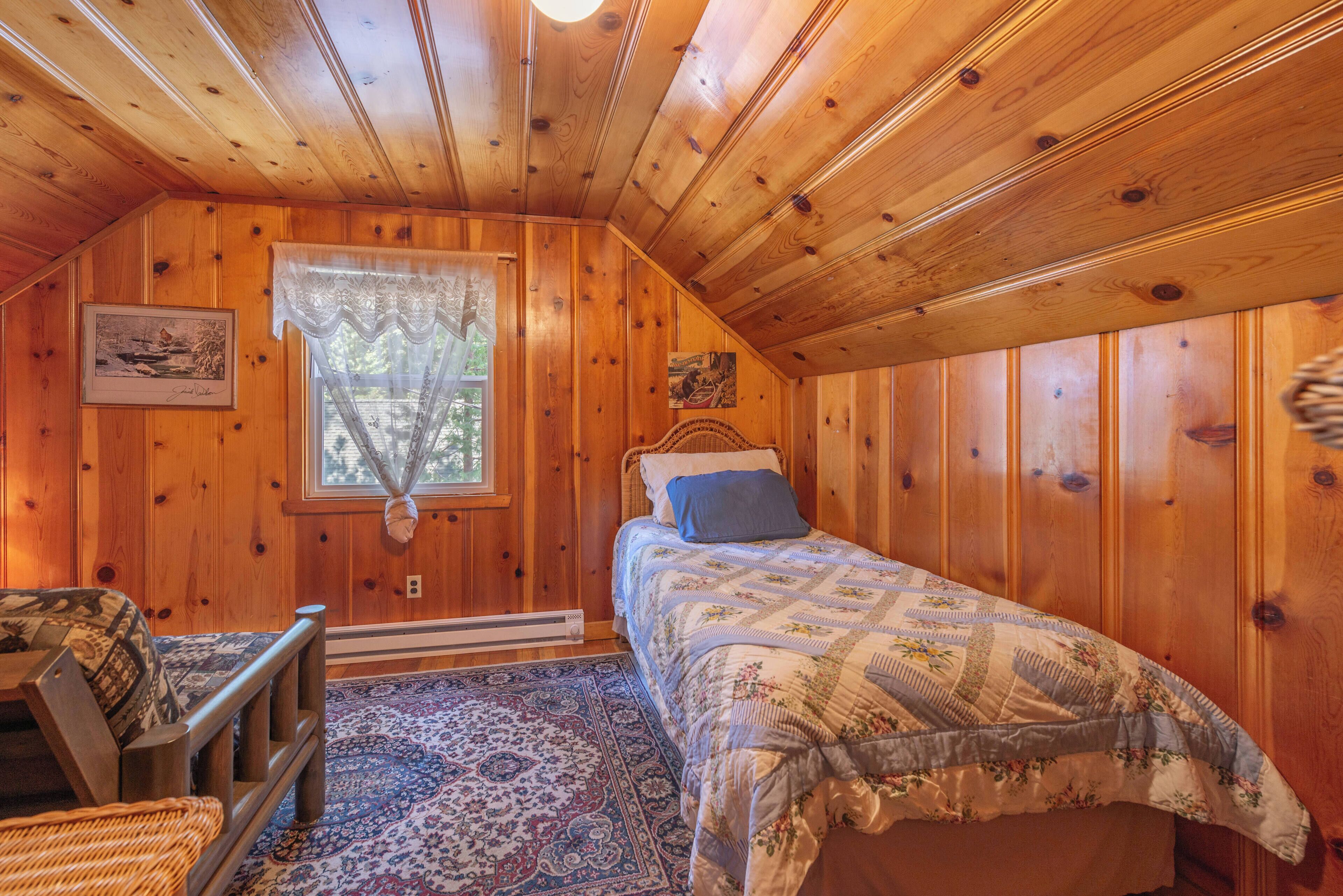 A cozy cabin bedroom with wooden walls, a bed with a floral comforter, a window with lace curtains, and a rustic chair.