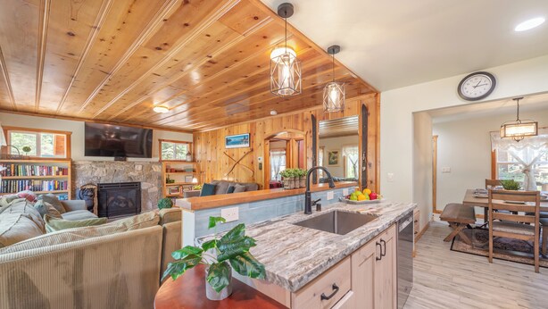 A spacious living area with a fireplace, wooden ceiling, and a stone fireplace surround. There is a kitchen with a large island, wooden cabinets, and a dining area with a table and chairs. - Walk to Beach and Town, Dogs OK, Cozy Fireplace, -Steelhead Mountain Retreat (Kings Beach)