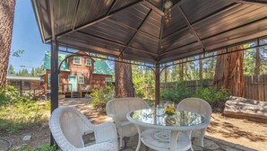 A covered patio area with wicker furniture and a glass table, surrounded by stone flooring and a wooden fence. - Walk to Beach and Town, Dogs OK, Cozy Fireplace, -Steelhead Mountain Retreat (Kings Beach)
