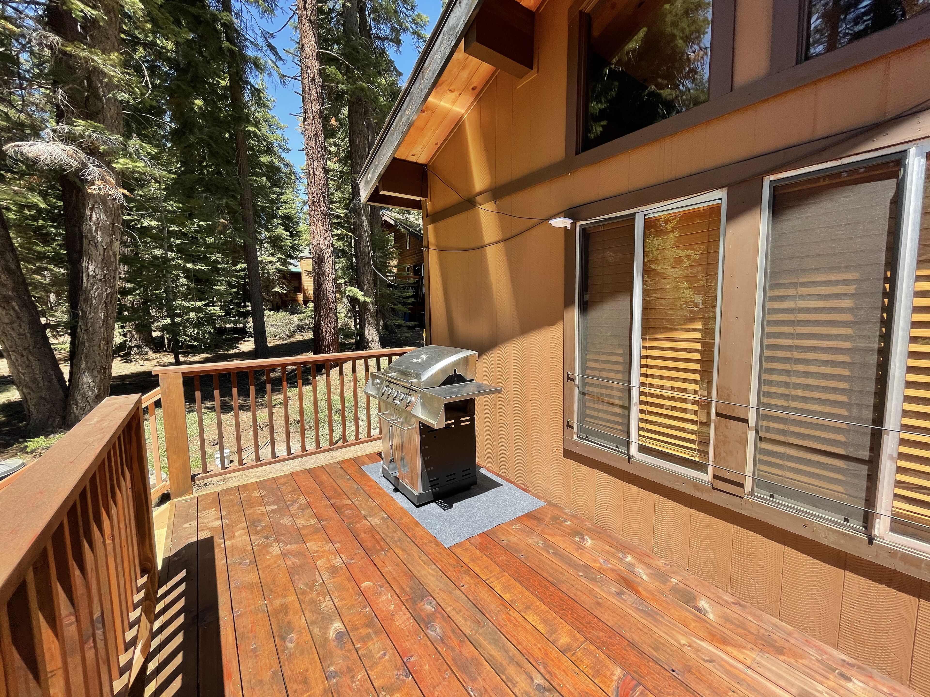 A wooden deck with a barbecue grill, large windows, and a view of tall trees.