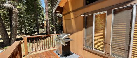 A wooden deck with a barbecue grill, large windows, and a view of tall trees.