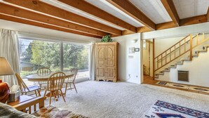 Ein gemütliches Zimmer mit Holzbalkendecke, einem Essbereich mit Stühlen und Tisch, einem großen Fenster mit Blick auf Bäume und einer Treppe mit dekorativer Teppich.
