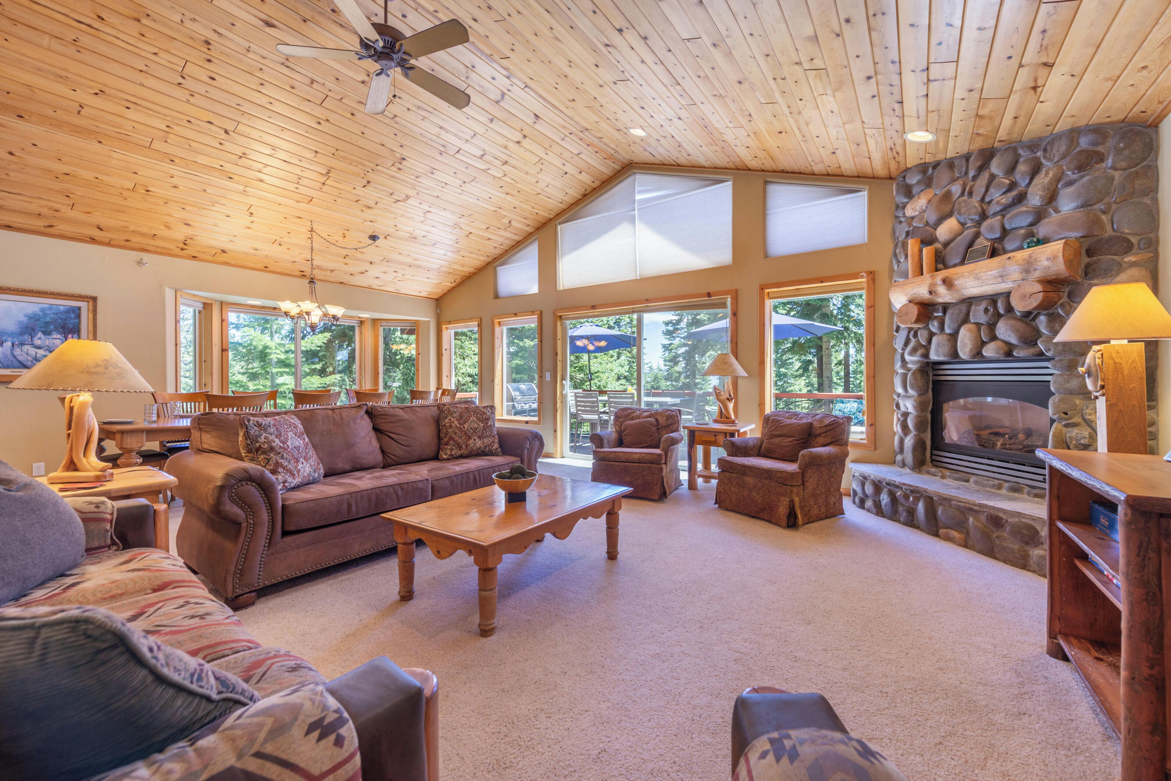 A spacious living room with a stone fireplace, wooden ceiling, and large windows.