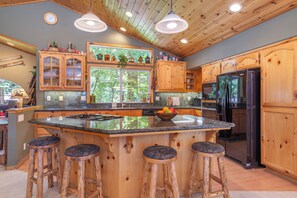 Una cocina con armarios de madera, una isla grande con taburetes, un refrigerador y una ventana con vistas a árboles.