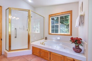 Un baño con ducha envuelta en vidrio, una bañera grande, armarios de madera y una ventana con vistas a árboles.