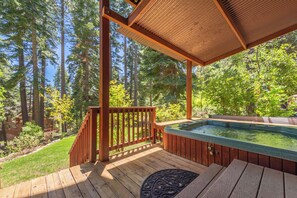 Una terraza de madera con un jacuzzi, rodeada de árboles altos y un cielo despejado.