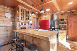 A wooden cabin interior with a kitchen area featuring a wooden bar, stools, a red refrigerator, and a stainless steel stove.