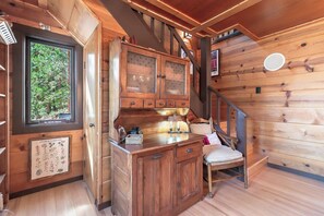 A wooden cabin interior with a wooden staircase, a cabinet with drawers, a small table with a lamp, and a window with a view of greenery.