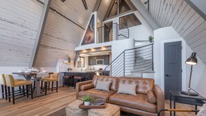 Un espace de vie de style loft avec un plafond haut, des poutres en bois, un canapé en cuir marron, une table à manger avec des chaises, et une cuisine en arrière-plan.