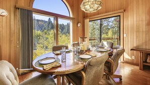 A dining area with a large table set for four, surrounded by wicker chairs, large windows offering a view of a forest, and a hanging light fixture. - HOA  w/ Pools & Hot Tubs, Dogs Ok- Northstar On The Course (Truckee)