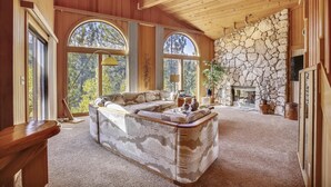 A cozy living room with a large sectional sofa, a stone fireplace, and large windows offering a view of trees. - HOA  w/ Pools & Hot Tubs, Dogs Ok- Northstar On The Course (Truckee)
