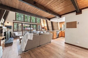 A spacious living area with a large sofa, wooden flooring, and a view of the outdoors through large windows. - Short Drive to the Beach, Hot Tub, Large Deck - All Decked Out (Carnelian Bay)