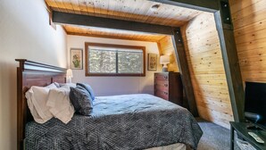 A bedroom with a sloped ceiling, a bed with a patterned bedspread, a dresser, a window with a view of trees, and a television on a stand. - Short Drive to the Beach, Hot Tub, Large Deck - All Decked Out (Carnelian Bay)