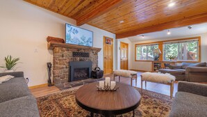 A cozy living room with a stone fireplace, a round wooden table with candles, and a large window with a view of trees. - Walk to Tahoe Park Private Beach, Hot Tub, Dogs OK- Cedar Crest (Tahoe City)