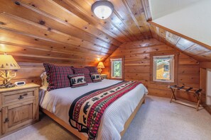 Ein gemütliches Schlafzimmer in einer Blockhütte mit Holzbalkendecke, einem Bett mit gemustertem Überwurf, einem Nachttisch mit Lampe und einem Fenster mit Blick auf Grünflächen.