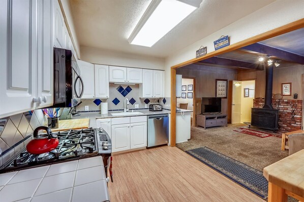 A compact kitchen with white cabinets, a red kettle, and a built-in stove. A dining area with a wooden table and chairs is visible through an open doorway.