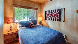 A bedroom with a large bed, two bedside lamps, a wooden nightstand, a window with blinds, and a wall-mounted tapestry.