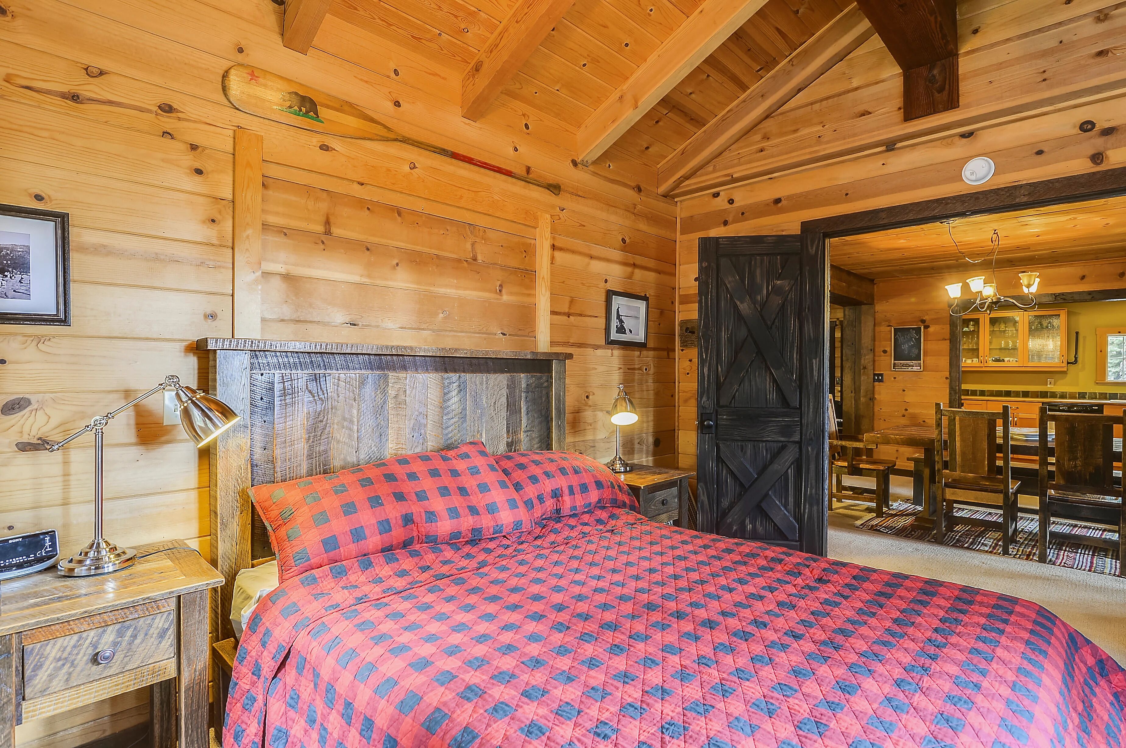 Un dormitorio de cabaña de madera con cama, lámparas de noche, cómoda y puerta que da a un comedor.