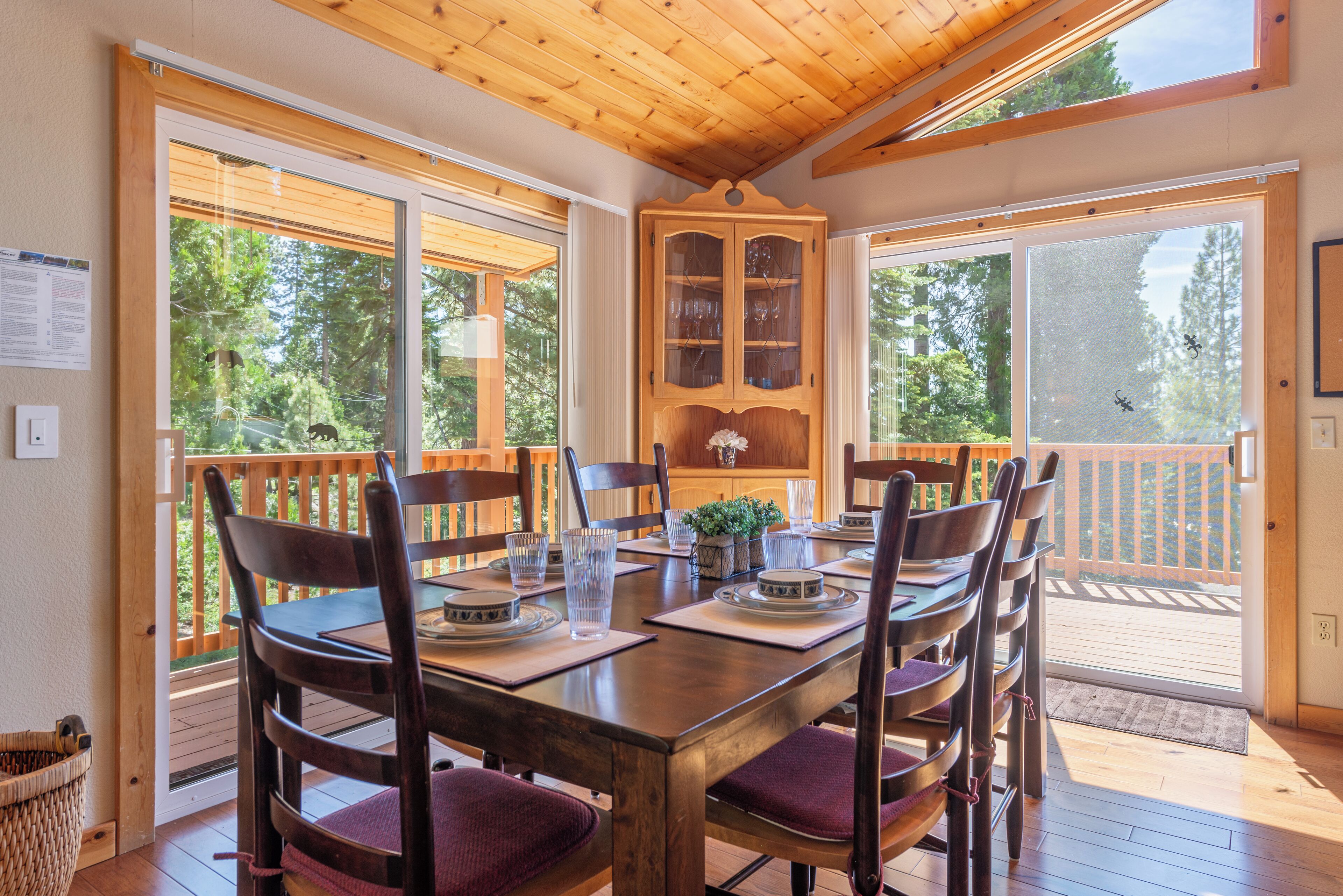 A wooden dining table with chairs, a cabinet with glass doors, and large windows with a view of trees.