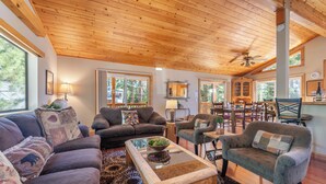 A cozy living room with a wooden ceiling, large windows, and a dining area with chairs and a table. - Overlook Cabin, Good View, Walk to Tahoe City, Wood Stove (Tahoe City)