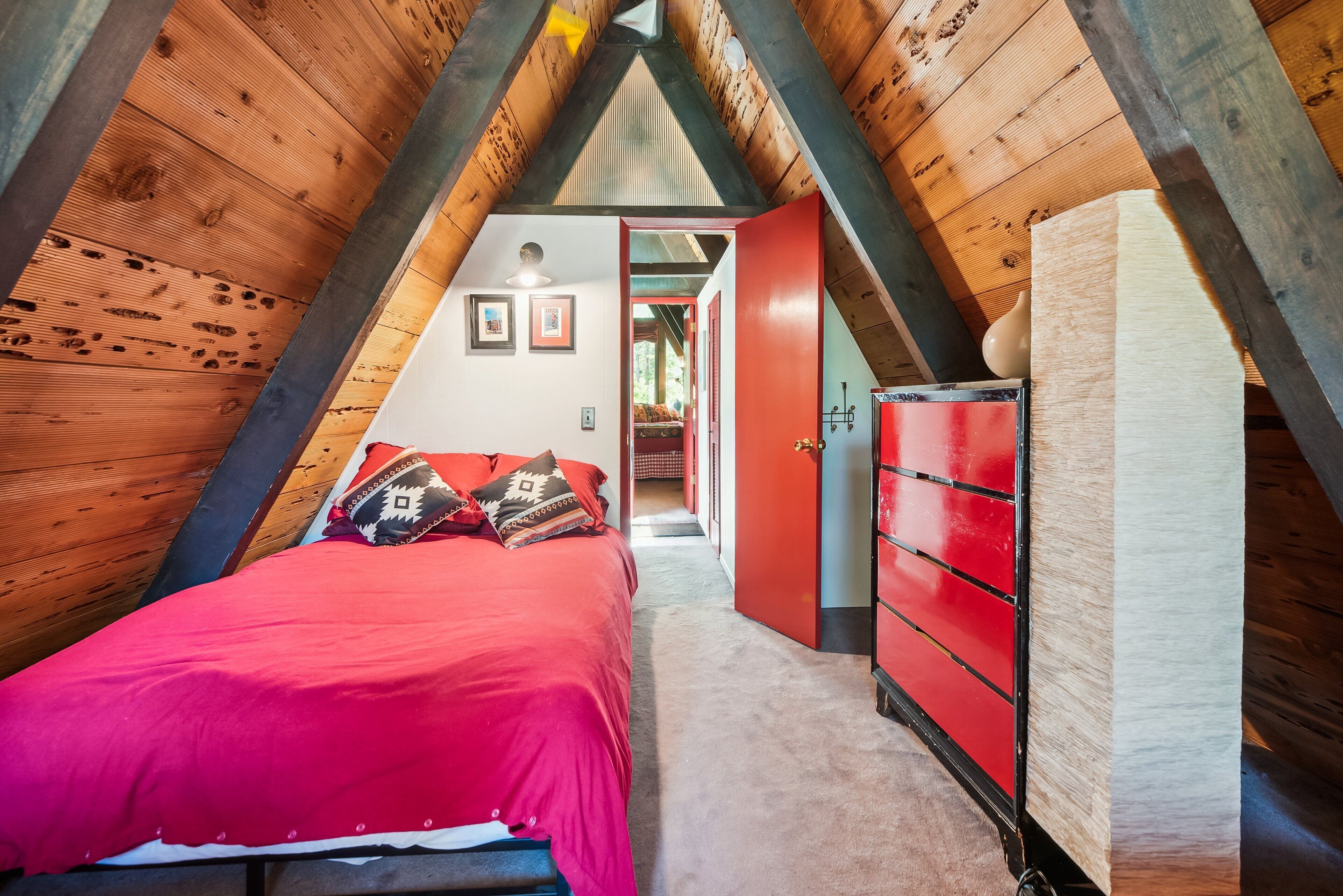 Une chambre avec un plafond en pente, un lit avec une literie rouge, une commode rouge et un couloir menant à une autre pièce.