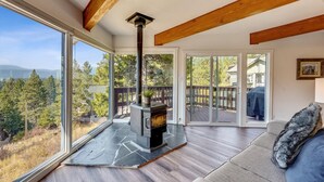 Una sala de estar acogedora con chimenea de piedra, ventanas amplias y vistas a una terraza y árboles.