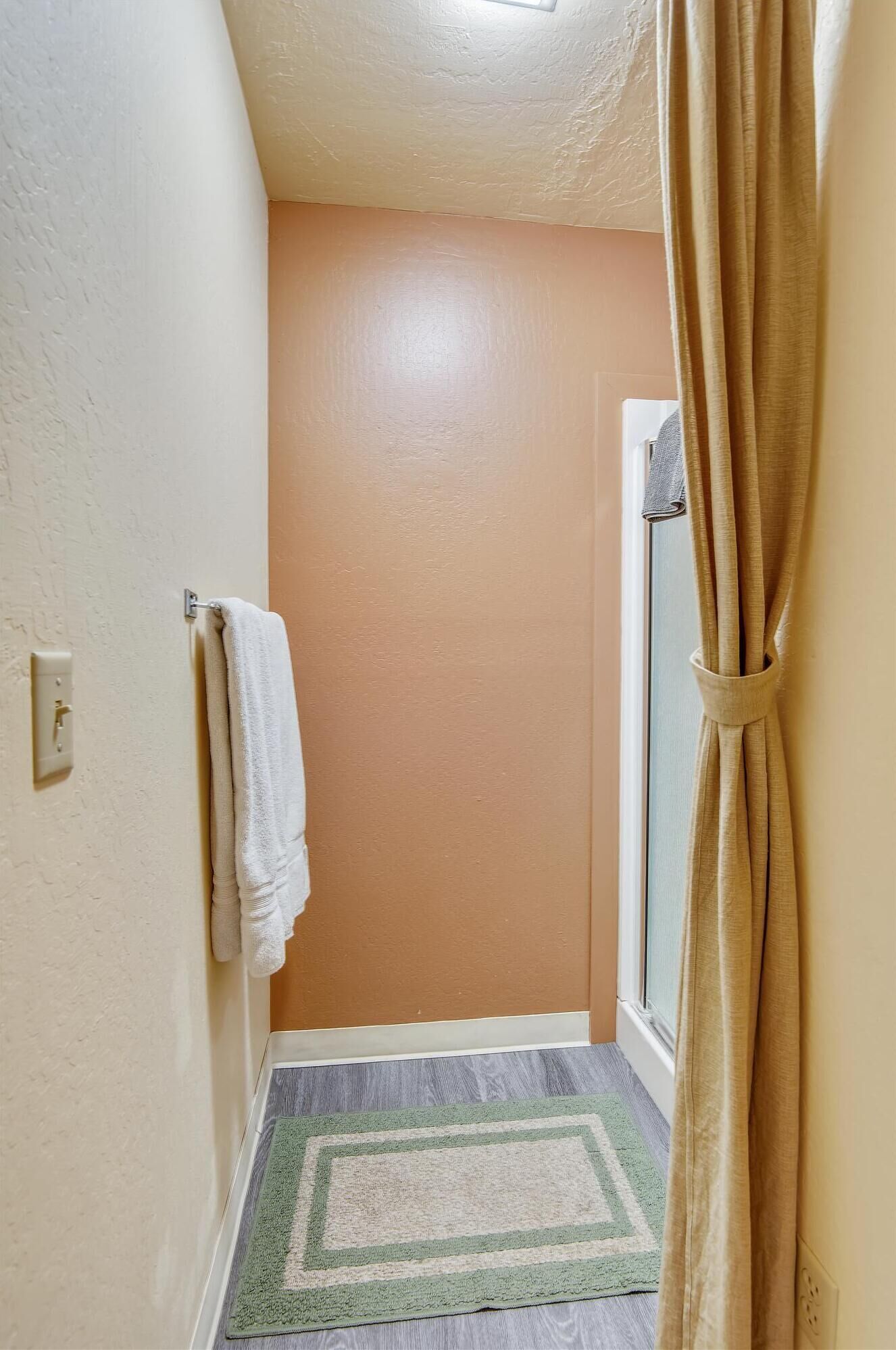 Ein schmales Dielenkorridor mit einer beigen Wand, einem weißen Handtuch an einem Haken, einem beigen Vorhang und einem gemusterten Teppich auf dem Boden.