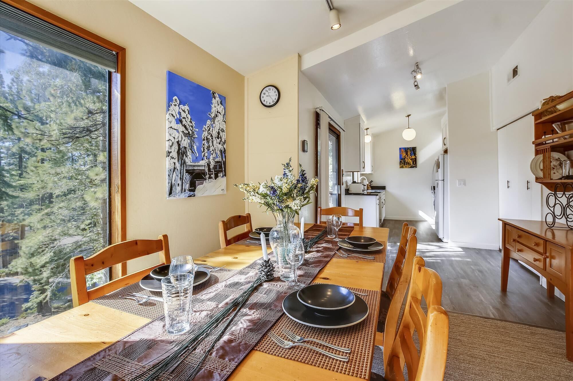 A dining area with a long wooden table set for four, a vase of flowers, and a painting on the wall.