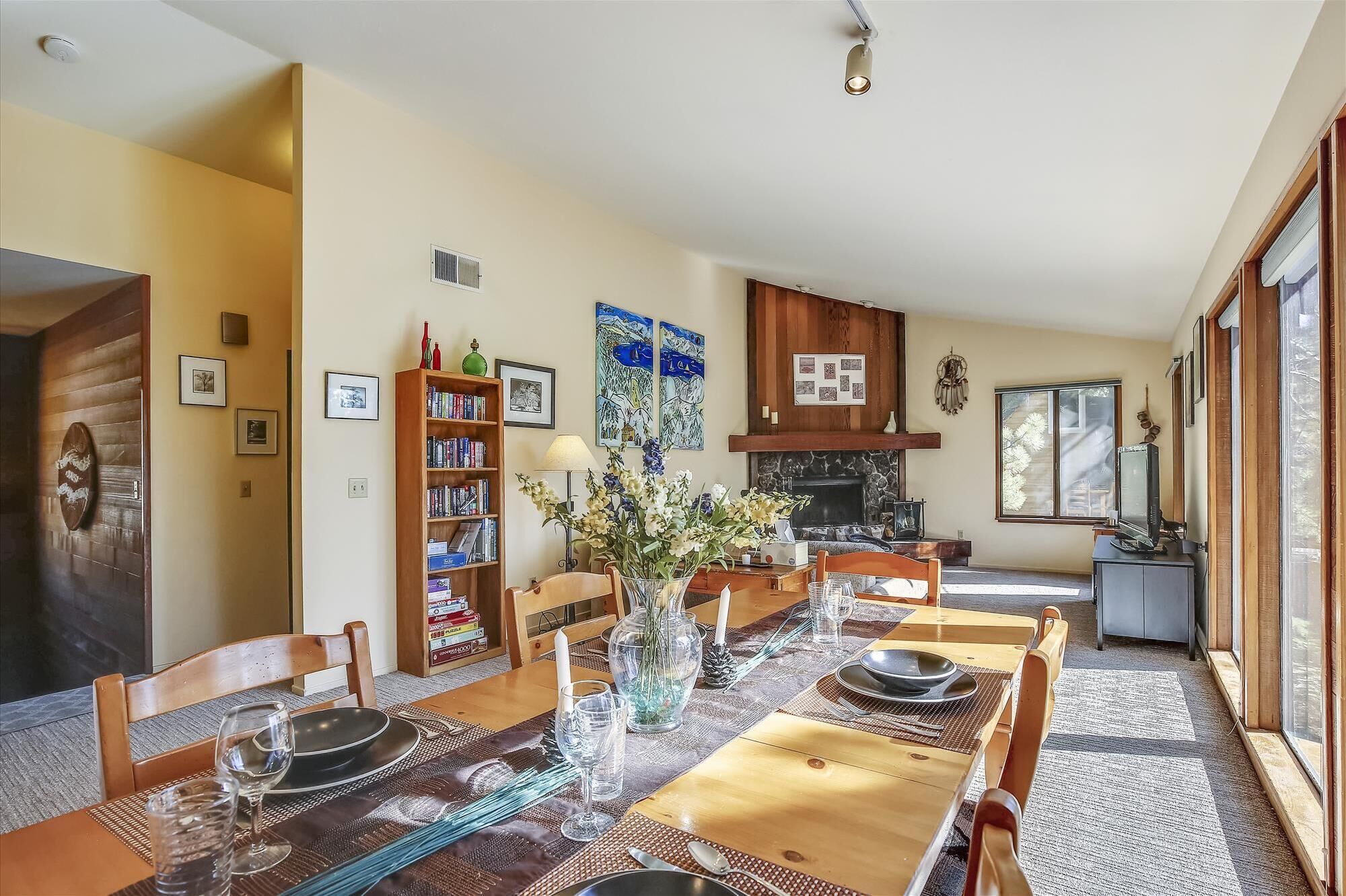 A dining area with a wooden table set for four, surrounded by chairs. There is a bookshelf filled with books and decorative items, a fireplace, and a television on a stand.