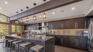 Een moderne keuken met donkere kastjes, een groot eiland met krukken, hangende verlichting en uitzicht op bomen door grote ramen.