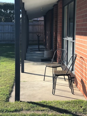 Ein überdachter Patio-Bereich mit Betonboden, zwei Metallstühlen, einem kleinen Tisch und einer hängenden Leuchte.
