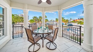 A covered outdoor area with a round table and chairs, a ceiling fan, and a view of a street with palm trees and buildings. - Siesta Breeze Retreat | Luxury Condo on SK w/ Heated Pool, Walking Distance to Beach & Village! (Siesta Key)