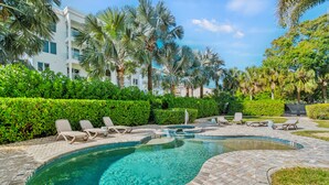 A swimming pool with lounge chairs, surrounded by palm trees and hedges, with a building in the background. - Siesta Breeze Retreat | Luxury Condo on SK w/ Heated Pool, Walking Distance to Beach & Village! (Siesta Key)