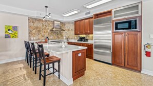 A modern kitchen with a central island, wooden cabinets, stainless steel appliances, and a stone backsplash. - Siesta Breeze Retreat | Luxury Condo on SK w/ Heated Pool, Walking Distance to Beach & Village! (Siesta Key)
