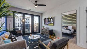 Una sala moderna con un sofá, una mesa de centro y un televisor. Hay un balcón con vistas a edificios.