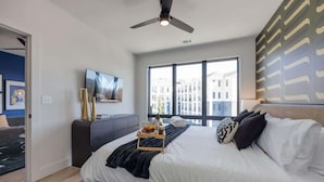Una habitación de hotel moderna con una cama grande, un ventilador de techo, una ventana con vistas a la ciudad y una mesita de noche con una bandeja de objetos.