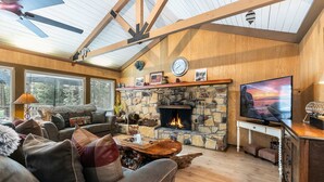A cozy living room with a stone fireplace, wooden beams, a ceiling fan, and a television.