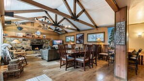 A spacious cabin interior with a stone fireplace, wooden dining table, and a large window overlooking a scenic landscape.