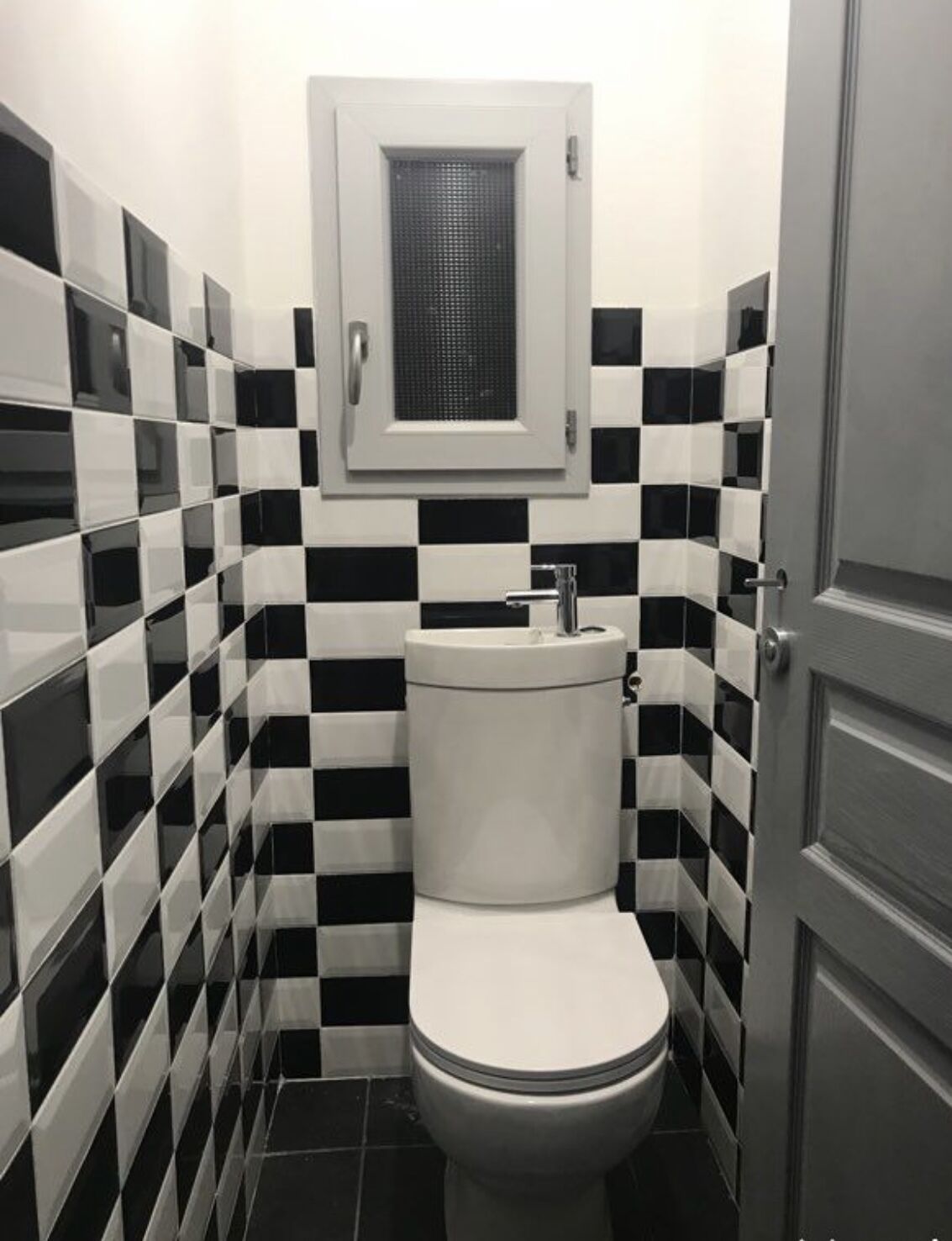 Et lille, kompakt badeværelse med toilet, håndvask og et vindue over håndvasken.